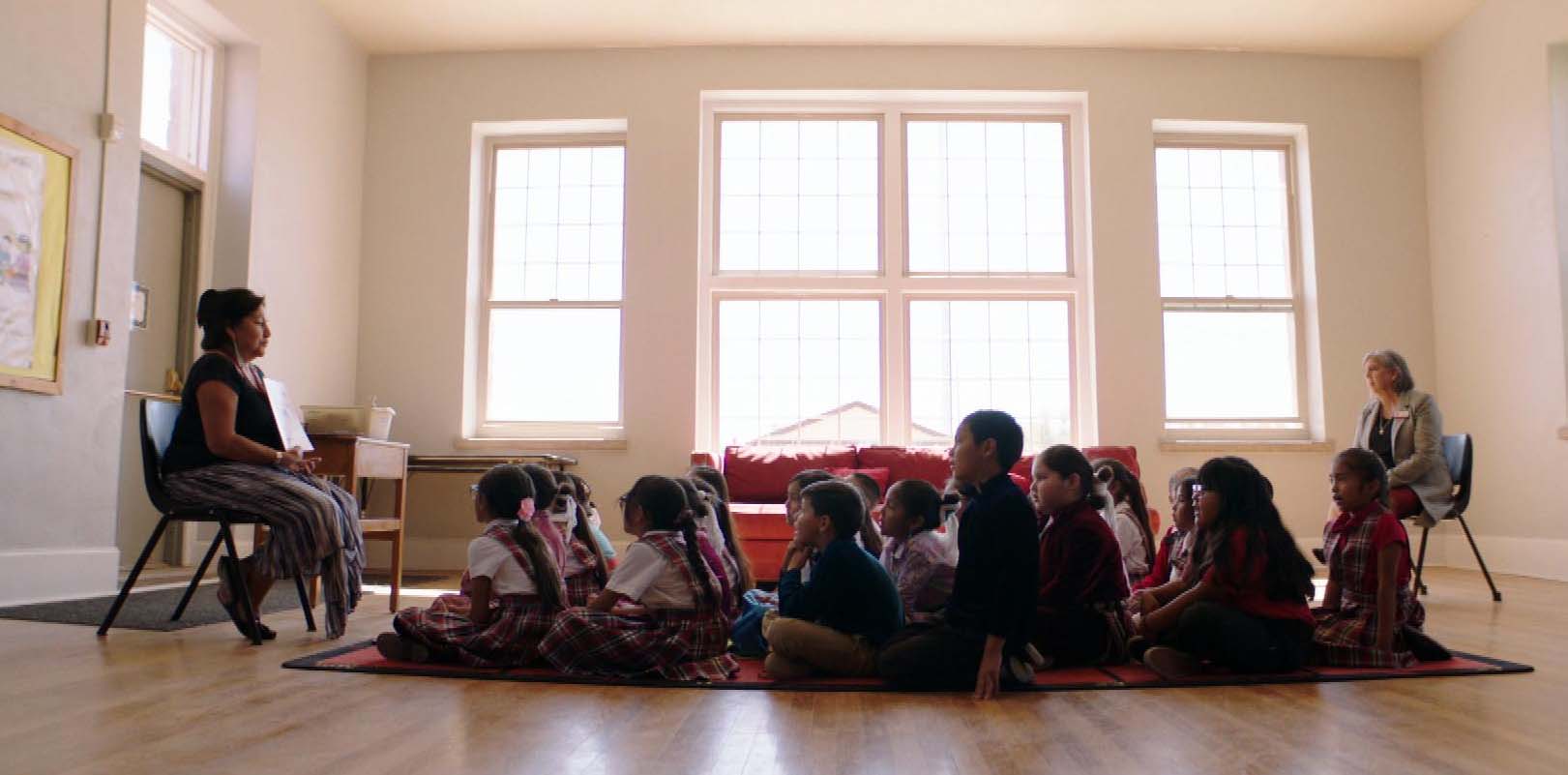 Students sit on the floor of a bright classroom looking up at a teacher who is holding open a book. Play video.