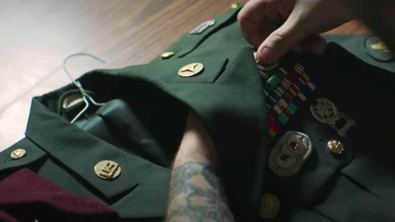 A close-up shows a hand adjusting ribbon racks and badges on a dark green military dress uniform jacket lying on a wooden surface next to a maroon beret.