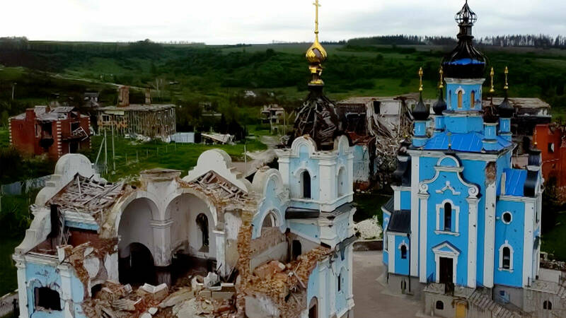An aerial view of two churches, one blue and white, the other white and light blue, standing amongst the ruins of other buildings. The white and light blue church is heavily damaged with its roof caved in and rubble surrounding its base. The blue and white church appears intact with a bright blue tarp covering sections of its roof.