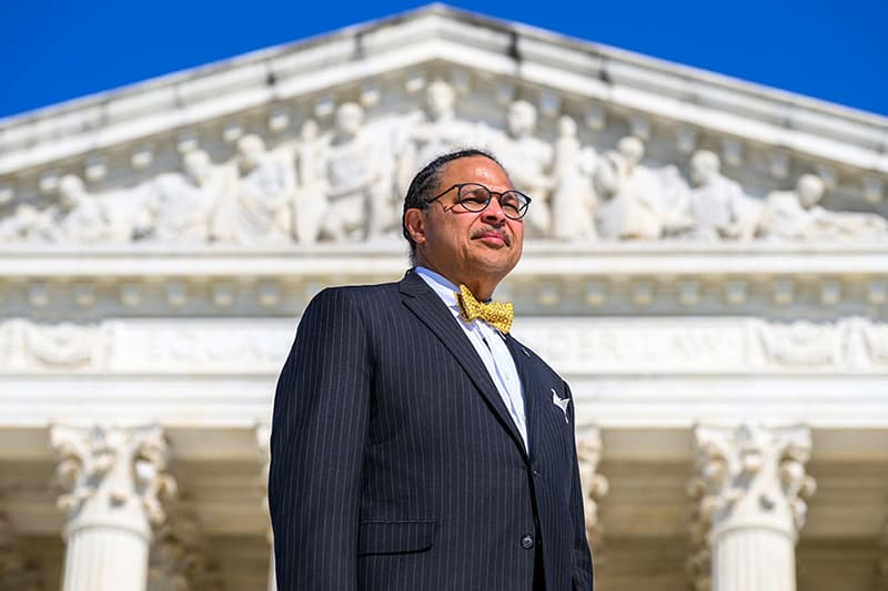 Marcus Cole, Dean of Notre Dame Law School, in a pinstripe suit and yellow bow tie, stands on the steps in front of the Supreme Court Building.
