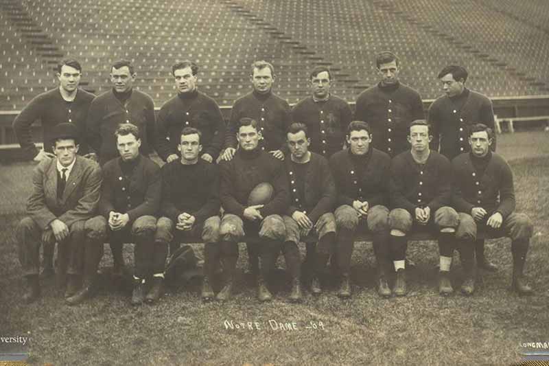 An old black and white photo of the 1904 Notre Dame football team posing in two rows on a bench with a stadium in the background. 