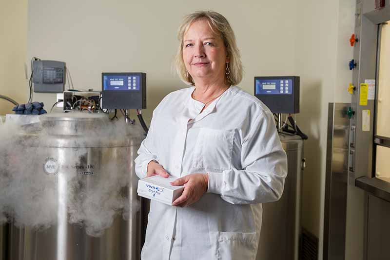 A researcher in a white lab coat stands in a lab, holding a small box, with two large cryogenic freezers behind her.