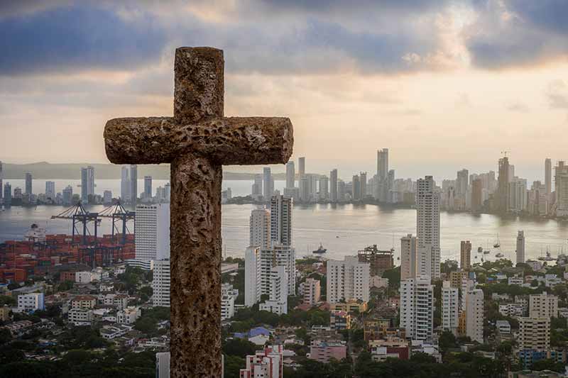A large stone cross in the foreground overlooks a busy port city with a bay and a skyline of modern buildings.