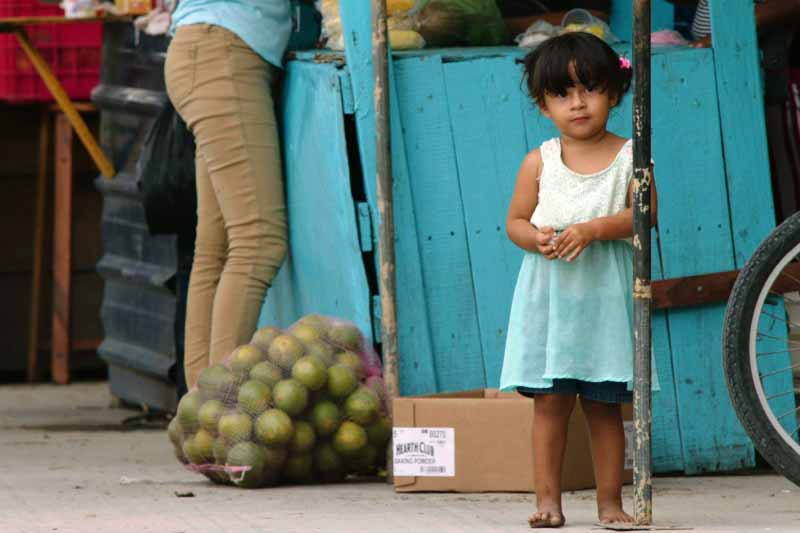 A small child with a concerned expression peeks out from behind a bright blue food stand.