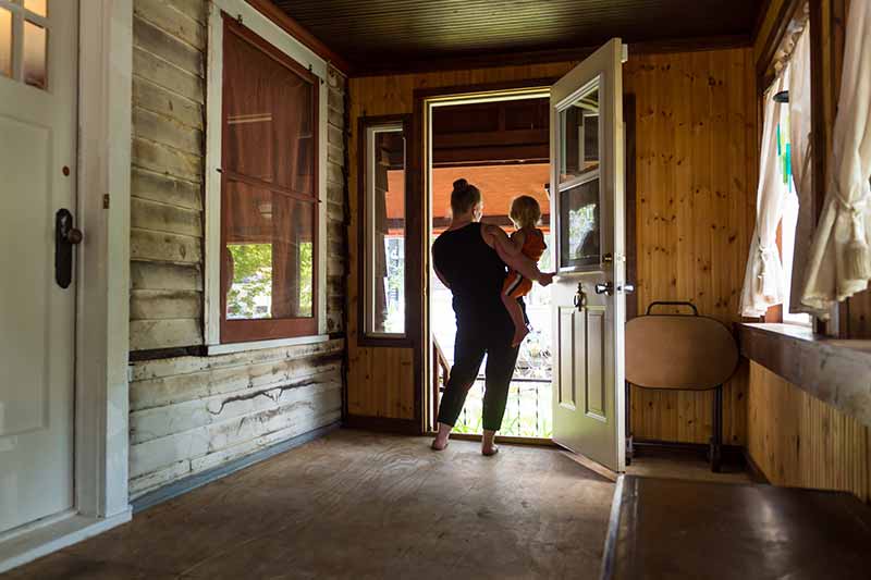 A woman holding a toddler opens a screen door to leave a rustic porch.