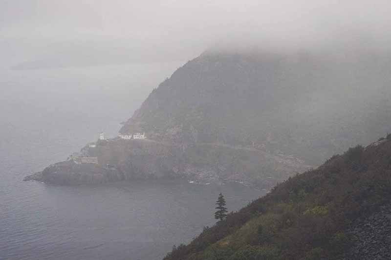 A lighthouse and buildings are built into the side of a cliff overlooking the ocean on a foggy day. 
