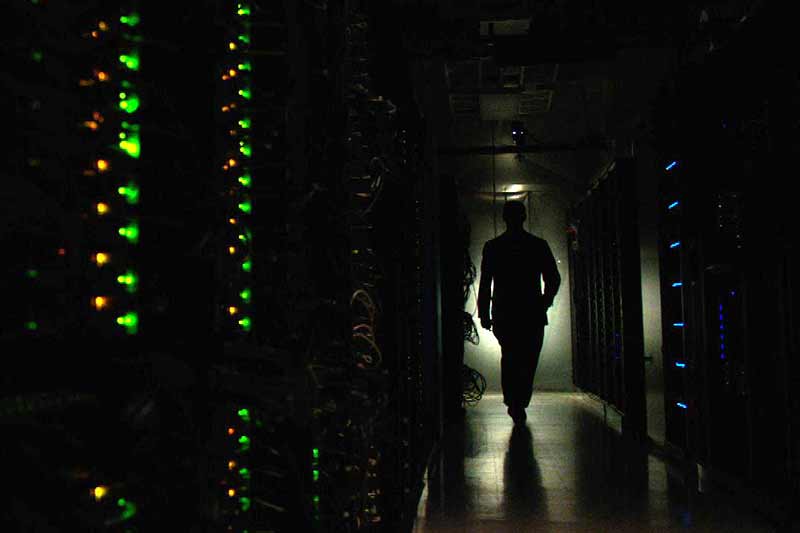A person is silhouetted as they walk down a dark hallway between computer servers with blinking lights. 