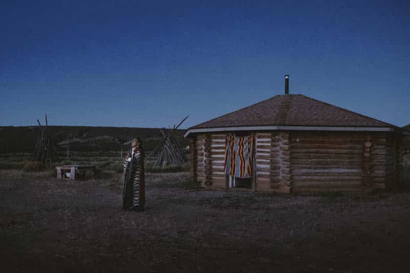 A person in traditional clothing stands in front of a hexagonal log cabin at dusk. 