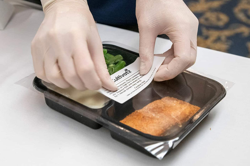 A person wearing gloves places a small white label on a prepackaged meal of salmon, mashed potatoes, and green beans.