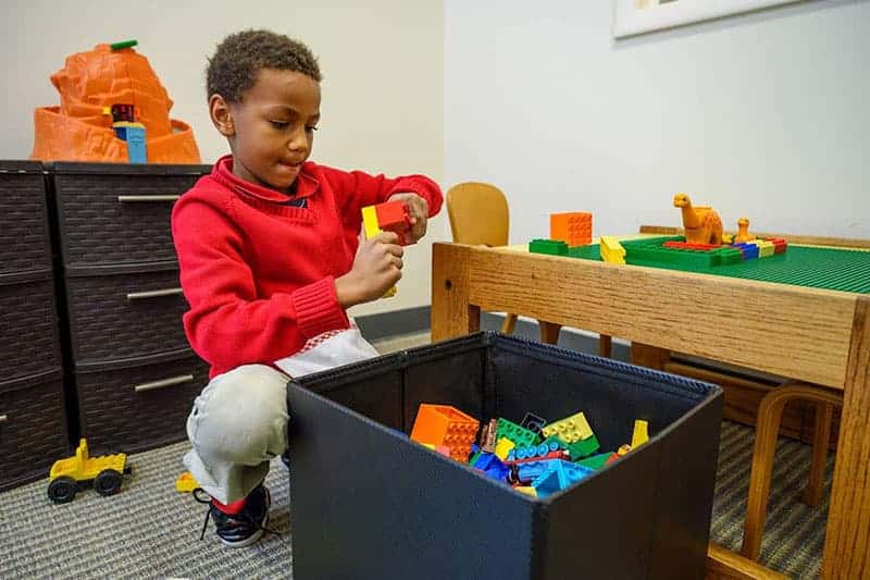 A young Black boy in a red sweater plays with yellow and red toy blocks from a black bin. 