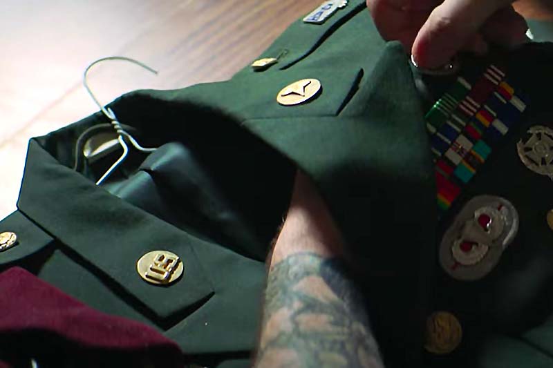 A person with a tattooed arm holds a military uniform jacket with service ribbons and a rank insignia on the collar. 
