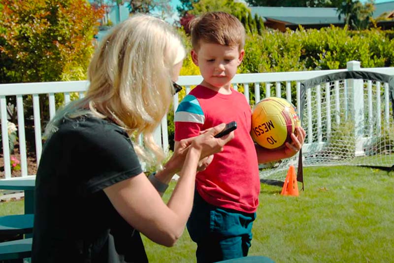 A woman and a young boy stand outside, looking down at a cell phone. The boy holds a soccer ball.