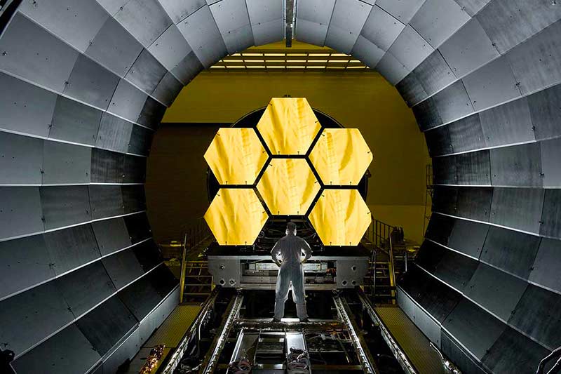 A man stands inside a large arch, looking at a hexagonal gold mirror, possibly from the James Webb Space Telescope.