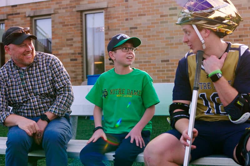 A group of three people sitting on a bench outdoors. A young boy in the center is wearing a green Notre Dame Men's Lacrosse t-shirt. He is flanked by an adult man on the left and a young man in a lacrosse uniform and helmet on the right.