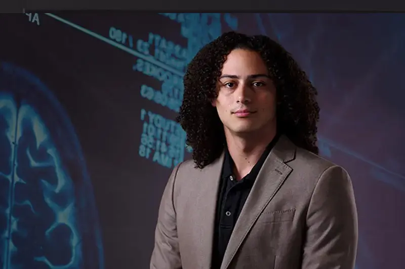 A headshot of a person with shoulder-length curly hair wearing a gray blazer and black shirt. They stand in front of a blurred image of a brain scan with medical data.