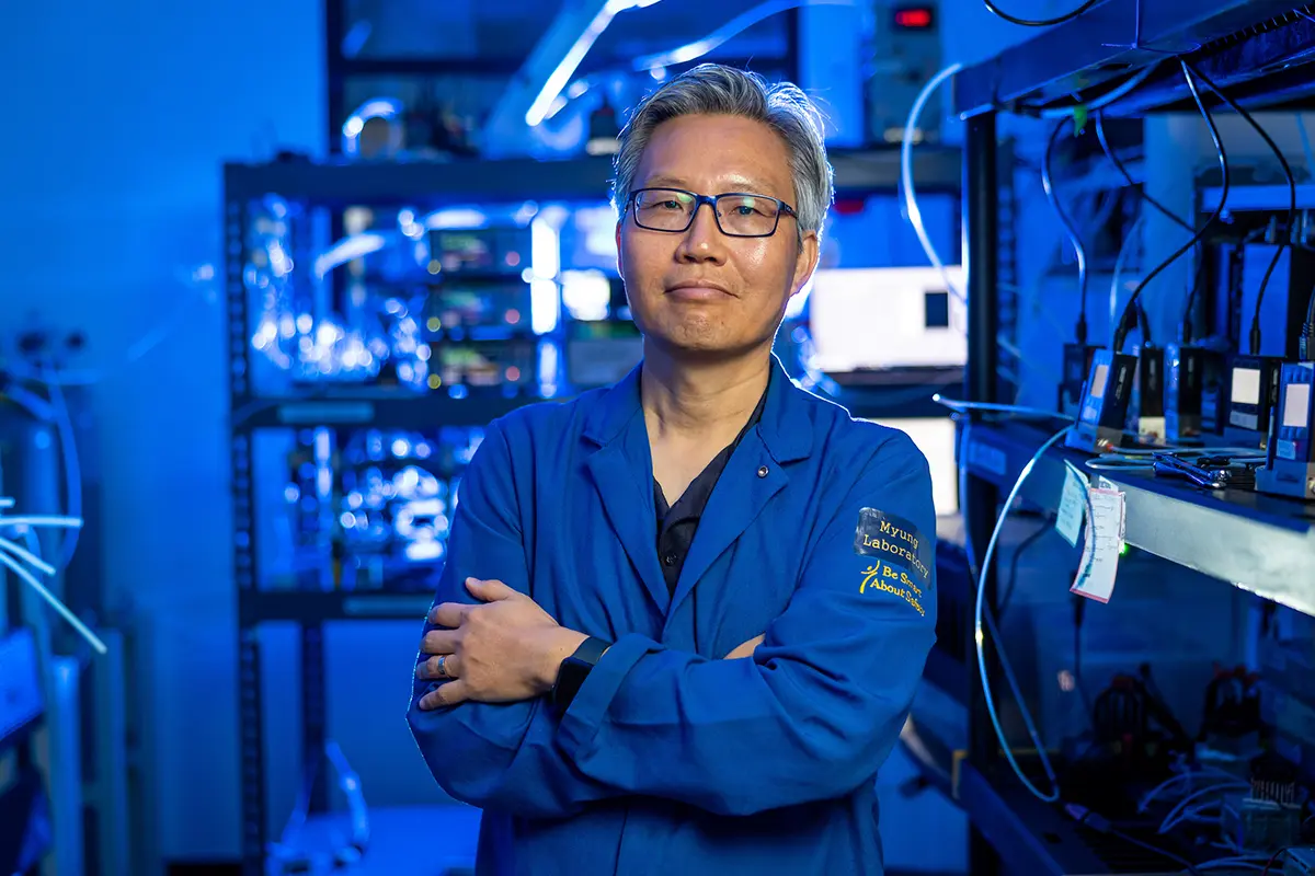 A scientist with gray hair and glasses, wearing a blue lab coat with a 'Myung Laboratory' patch, stands with his arms crossed in a brightly lit blue laboratory setting.