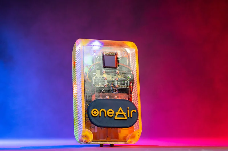 A translucent amber electronic device, revealing a circuit board with components and a small dark display. A black oval badge with orange “oneAir” text, where the A is a triangle, is on the front. It rests against a blue-to-red gradient background.