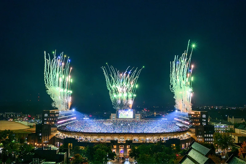 Notre Dame Stadium at night, packed with spectators, illuminated by bright stadium lights and green and white fireworks exploding above the arena.