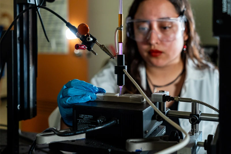 A student wearing safety glasses and blue gloves uses a scientific instrument emitting a purple plasma beam.