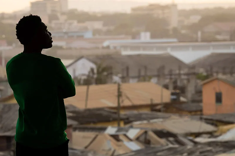 Silhouette of a person in a green shirt with arms crossed looking out over a densely built, hazy cityscape.