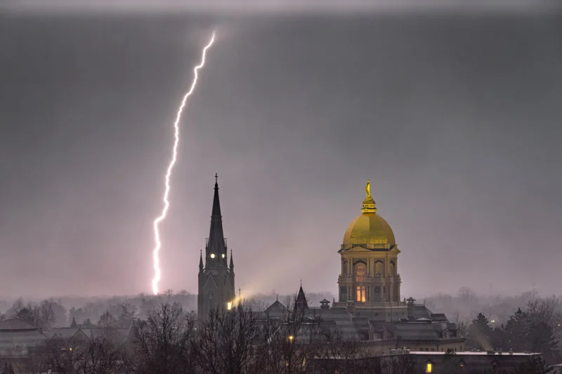 A bright lightning bolt flashes down behind the dark spire of the Basilica of the Sacred Heart and the illuminated Golden Dome of Notre Dame's Main Building under a grey, stormy sky.