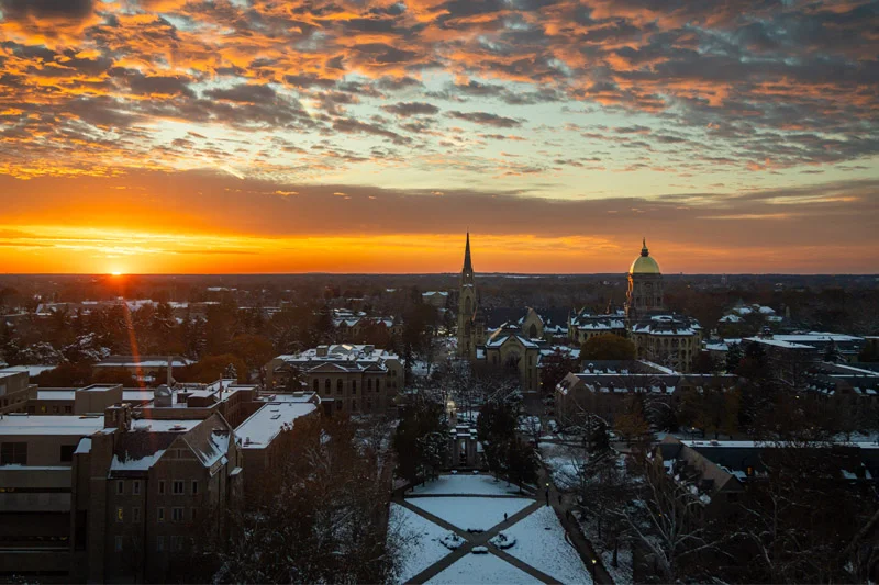 Aerial view of the University of Notre Dame campus covered in snow at sunset. The Golden Dome and Basilica of the Sacred Heart spire stand against a vibrant orange sky above snow-dusted rooftops and a central quad with intersecting walkways.