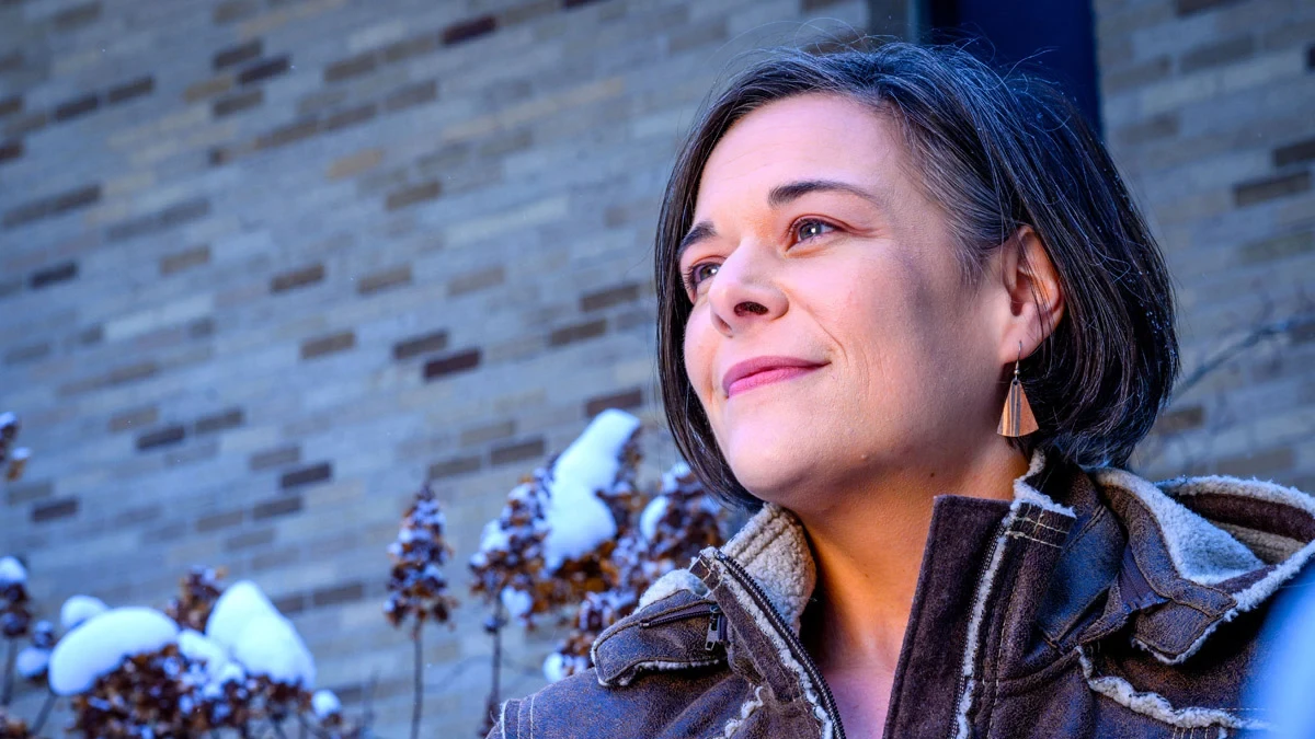 A woman with short dark hair smiles slightly while looking off-camera. She wears a brown, fleece-lined winter jacket. In the background, snow rests on dried plants in front of a tan brick wall.