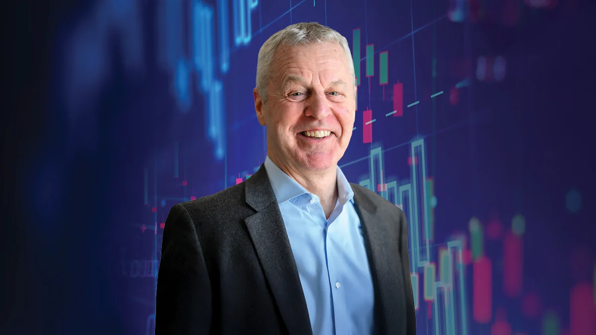 Smiling older man with short gray hair wearing a gray blazer and light blue shirt, set against a deep blue background overlaid with colorful financial candlestick chart graphics in teal, pink, and red.