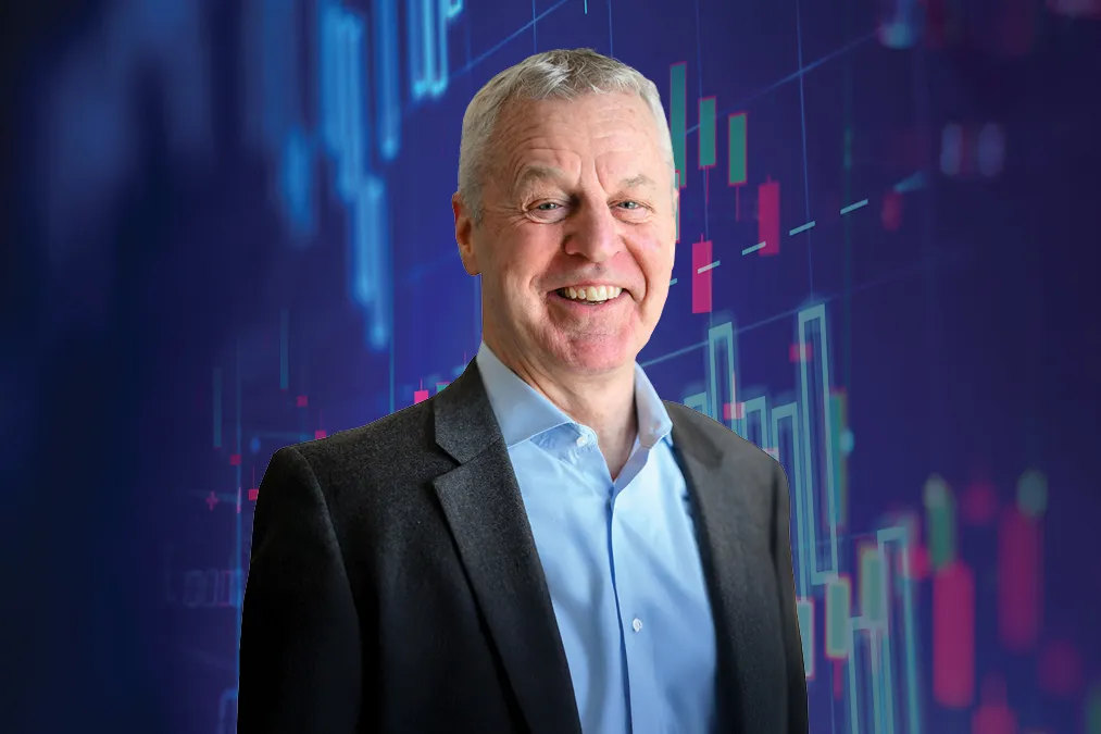 Smiling older man with short gray hair wearing a gray blazer and light blue shirt, set against a deep blue background overlaid with colorful financial candlestick chart graphics in teal, pink, and red.