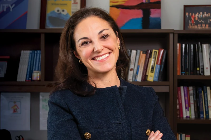 Broadly smiling woman with dark hair in a navy jacket with gold buttons, arms crossed, in front of bookshelves.