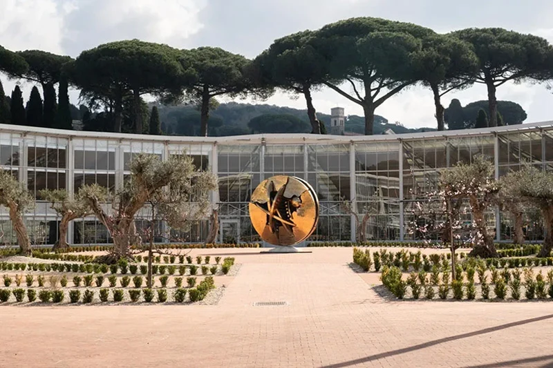 The bronze sculpture Desert Rose by Arnaldo Pomodoro is placed at the center of the ecological greenhouse at the Gardens of Castel Gandolfo. The curved glass structure—covered with solar panels—frames the work within a light-filled, sustainable environment. Stone pathways and carefully trimmed hedges unfold beneath a canopy of Mediterranean pines.