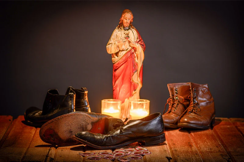 Statue of Sacred Heart of Jesus, two lit candles, surrounded by worn black and brown shoes and scattered red tokens on wood.
