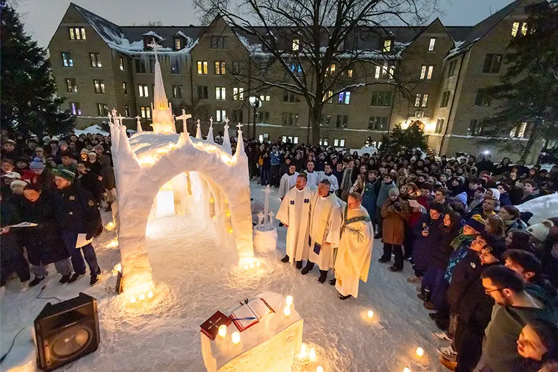 Students and three priests in white vestments gather for a night mass around a lit snow chapel and altar, snow-covered Sorin Hall in background.