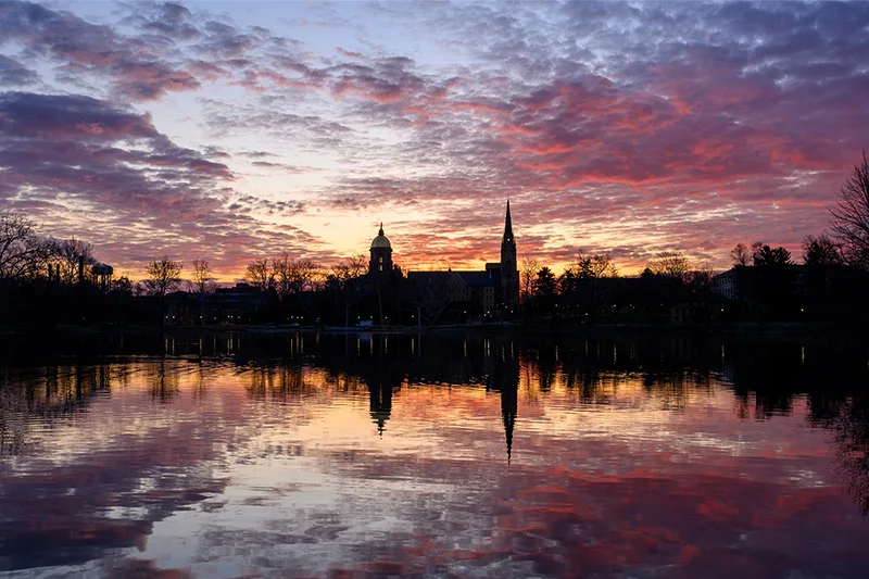 Golden Dome and Basilica of the Sacred Heart silhouetted against a red and orange sunset, reflected in a Notre Dame lake.