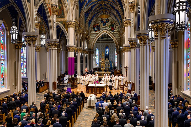 Congregation and clergy gather at the Basilica of the Sacred Heart with a purple banner and white-draped coffin.