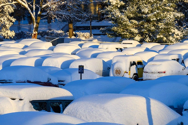A snow-covered parking lot at the University of Notre Dame. Dozens of cars are buried under thick mounds of white snow, with one yellow Jeep featuring an ND logo on the spare tire cover visible amidst the winter landscape.