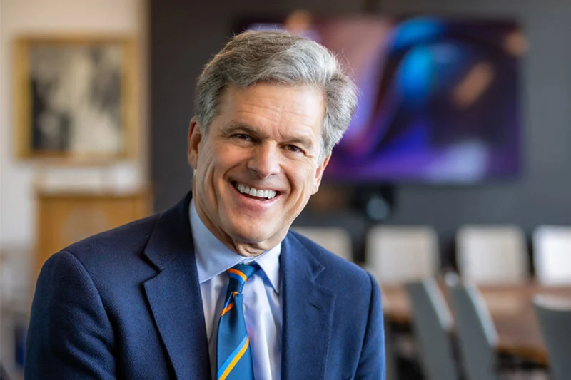 Smiling man with gray hair wearing a navy blazer, light blue dress shirt, and blue striped tie with orange accents, seated in a conference room with artwork and a monitor visible in the blurred background.
