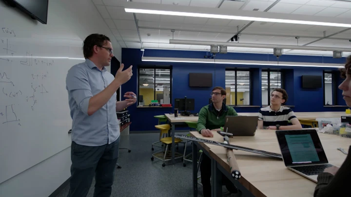 A man in a light blue shirt gestures toward a whiteboard covered in equations while instructing two students in a campus lab. The students sit at a wooden table with a laptop and a weather measuring drone against a blue wall.