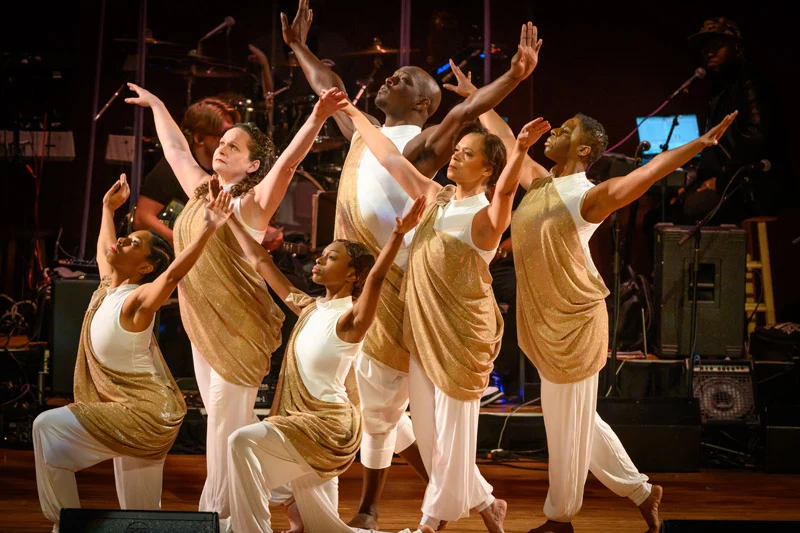 Six dancers in white outfits with shimmering gold wraps perform a choreographed routine on a stage. They are posed with arms extended and gazes lifted upward during a live performance with a band in the background.