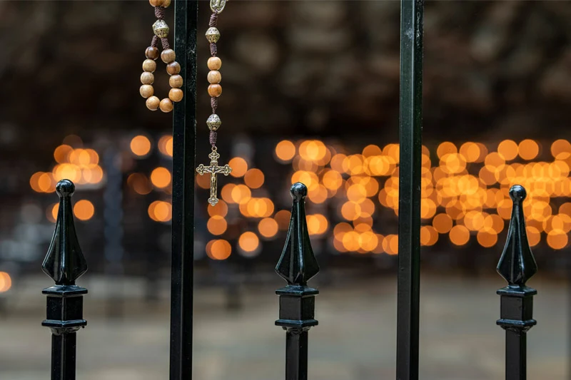 A wooden rosary with a silver crucifix hangs from a black iron fence. In the soft-focus background, dozens of glowing candles create a warm, orange bokeh effect at the Grotto of Our Lady of Lourdes.