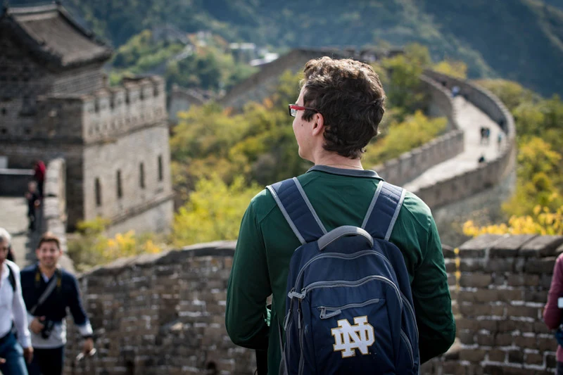 A student wearing a navy blue backpack with the gold Notre Dame ND logo looks out over the Great Wall of China. The stone fortifications wind through green, hilly terrain under a clear sky.