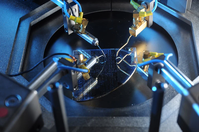 Precision robotic probes make contact with a blue-tinted microchip wafer during a technical inspection. The scene is illuminated with cool blue light, highlighting the intricate circuitry and metallic components.