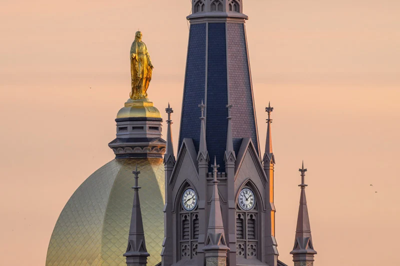 Close-up of the Golden Dome and the Basilica of the Sacred Heart at the University of Notre Dame against a soft sunset sky. The golden statue of Mary stands atop the dome next to the Basilica's gothic spires and clock face.
