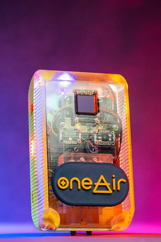 A translucent amber electronic device, revealing a circuit board with components and a small dark display. A black oval badge with orange “oneAir” text, where the A is a triangle, is on the front. It rests against a blue-to-red gradient background.