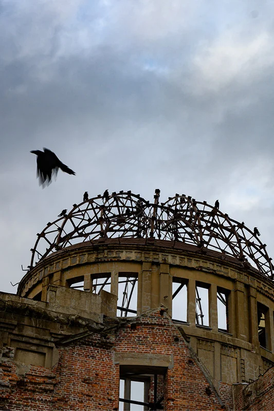 The ruined Hiroshima Peace Memorial (Genbaku Dome) featuring exposed brick and a skeletal metal dome against an overcast sky. Several dark crows perch on the dome, one flying past.