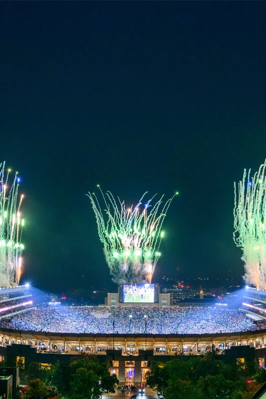 Notre Dame Stadium at night, packed with spectators, illuminated by bright stadium lights and green and white fireworks exploding above the arena.