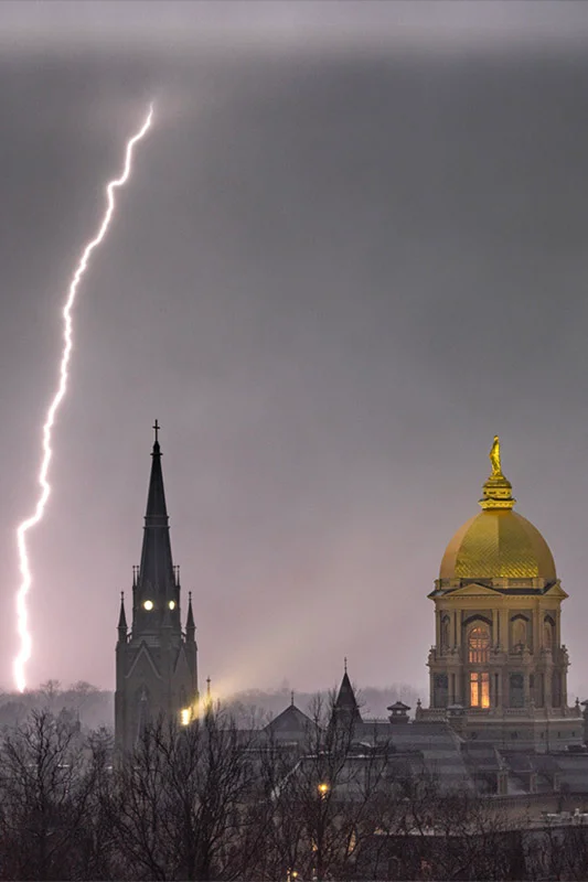 A bright lightning bolt flashes down behind the dark spire of the Basilica of the Sacred Heart and the illuminated Golden Dome of Notre Dame's Main Building under a grey, stormy sky.