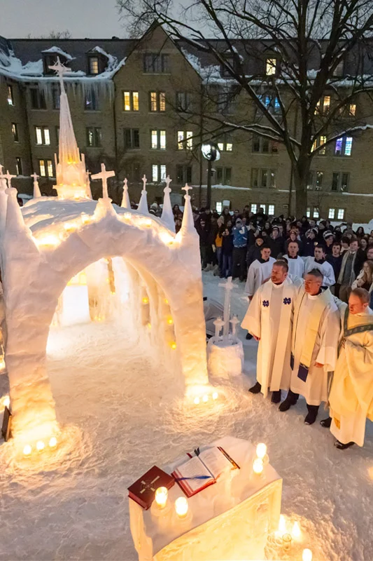 Students and three priests in white vestments gather for a night mass around a lit snow chapel and altar, snow-covered Sorin Hall in background.