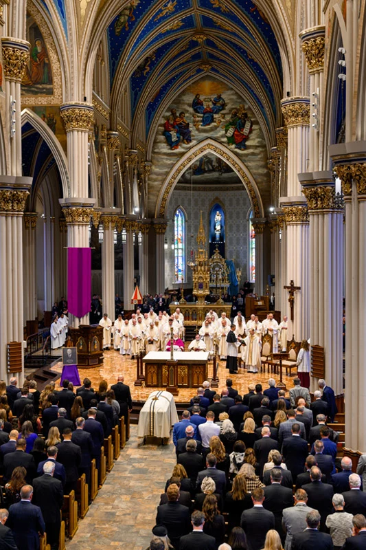 Congregation and clergy gather at the Basilica of the Sacred Heart with a purple banner and white-draped coffin.
