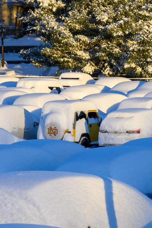 A snow-covered parking lot at the University of Notre Dame. Dozens of cars are buried under thick mounds of white snow, with one yellow Jeep featuring an ND logo on the spare tire cover visible amidst the winter landscape.